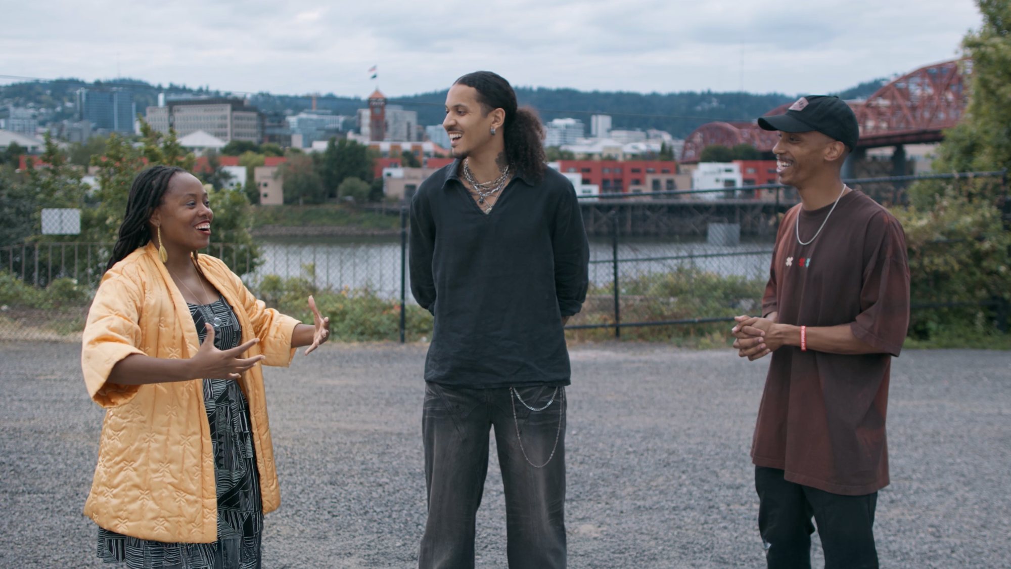 Three people talking and laughing on a gravel lot above the Willamette River, with the Portland skyline and Broadway Bridge behind them
