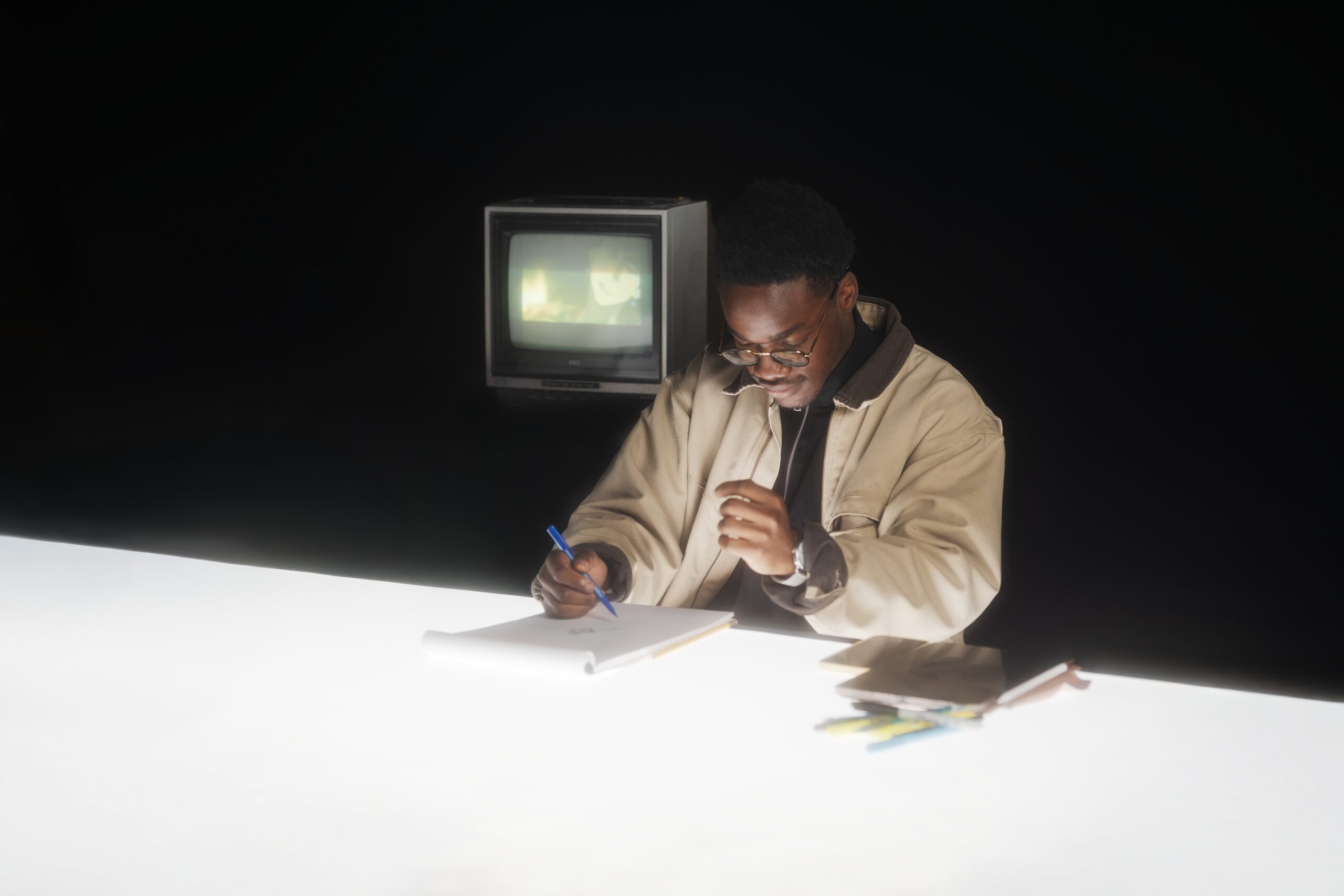 Subject writing at a desk beside a vintage TV glowing with static in a dark studio
