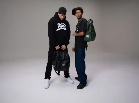 Two players in Vika apparel on a studio cyc, one in a black hoodie holding a backpack, the other with a green PDX Football backpack