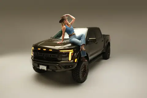 Model in a cowboy hat sitting on the hood of a black Ford Raptor with its amber lights on, studio cyc