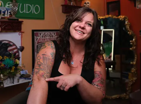 Smiling tattoo artist pointing to the tattoos on her arm inside her studio