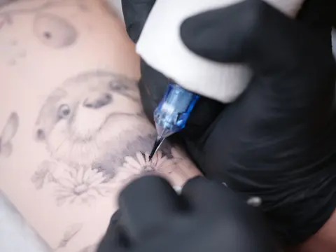 Close-up of a gloved hand tattooing a fine-line otter and daisies on skin