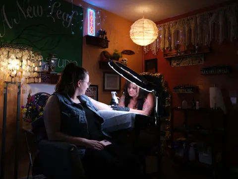 Wide view of an artist tattooing a client under a ring light in a warm, cluttered studio