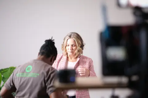 Crew member prepping talent in a pink tweed jacket beside the camera