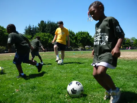 Kids in green camp jerseys dribbling soccer balls across a sunny park field
