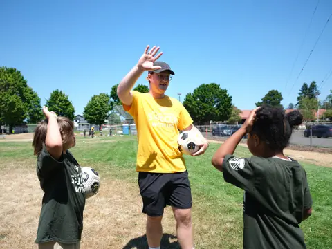 Coach in a yellow shirt high-fiving two young campers holding soccer balls