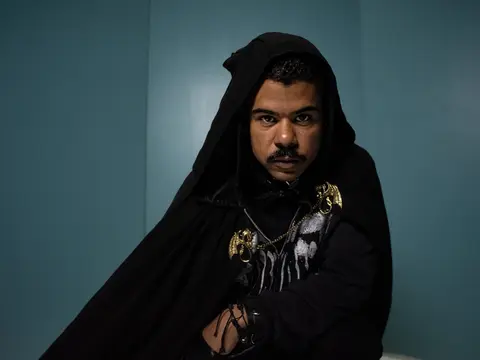 ILoveMakonnen in a black hooded cloak with gold chains against a teal wall